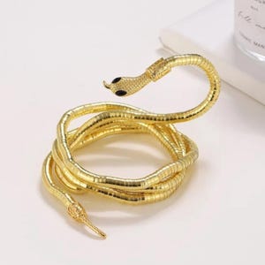 Puede incluir: Una pulsera de serpiente de metal dorado flexible con un diseño en espiral. La cabeza de la serpiente tiene ojos negros incrustados y escamas texturizadas, mientras que la cola es puntiaguda. La superficie metálica tiene un acabado pulido, creando una apariencia segmentada continua que se enrolla sobre sí misma en múltiples bucles.