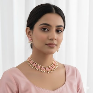 Puede incluir: Una mujer lleva un collar floral rosa y pendientes a juego. El collar y los pendientes presentan detalles en forma de flor rosa y pequeñas perlas blancas. Las joyas se lucen sobre un top rosa.