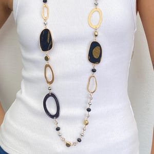 Puede incluir: Collar largo dorado con una variedad de formas de colgantes. El collar presenta formas abstractas negras y doradas, círculos abiertos dorados y pequeñas cuentas negras y doradas. El collar se lleva sobre una camiseta sin mangas blanca.