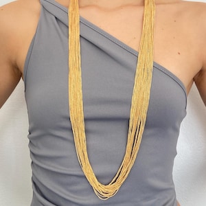 Peut inclure: Un long collier doré composé de multiples brins. Le collier est porté sur un haut gris, créant un effet de superposition. Les brins sont fins et semblent être en métal, reflétant la lumière. Un bijou élégant.
