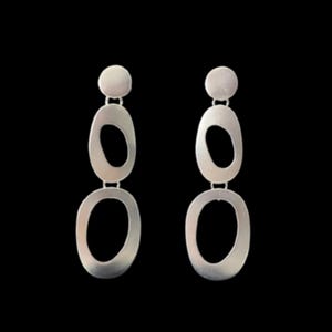 May include: A pair of silver-toned drop earrings featuring a vertical arrangement of three connected elements. Each earring consists of a small solid circular top piece followed by two larger open oval-shaped links with a smooth matte finish.