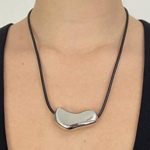 Op de afbeelding: Een ketting met een gladde, gepolijste zilverkleurige metalen hanger in een onregelmatige, organische, boonachtige vorm. De hanger hangt aan een eenvoudig, dun zwart koord dat om de nek rust, wat zorgt voor een strak en reflecterend uiterlijk.