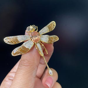 May include: A gold-toned dragonfly brooch with intricate detailing. The wings feature a brown and white pattern, while the body has a large peach-colored gemstone. The brooch is embellished with small, sparkling crystals.