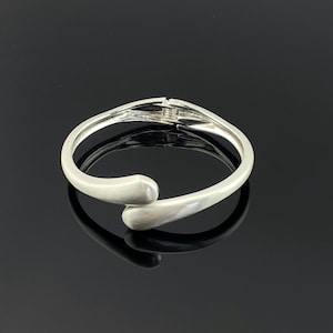 Puede incluir: Una pulsera plateada con un diseño curvo. La pulsera tiene un acabado liso y mate y un cierre de broche. Los extremos de la pulsera se curvan hacia adentro, creando un diseño abierto único. La pulsera tiene aproximadamente 6,35 cm de diámetro.