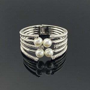 May include: A silver-toned hinged cuff bracelet featuring four parallel bands with a twisted rope texture. The open front design is accented by four round white faux pearls and small clear crystals embedded near the ends of each band.