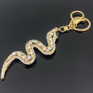 Pode incluir: Um porta-chaves de metal dourado com um pendente em forma de cobra coberto de strass iridescentes multicoloridos. A cobra está representada numa posição sinuosa em forma de S e está presa a um anel circular dourado e a um fecho lagosta.