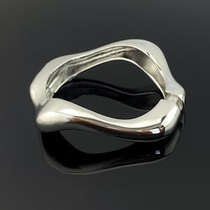 Puede incluir: Un brazalete rígido articulado de tono plateado pulido con una silueta ondulada, orgánica e irregular. La superficie metálica es lisa y reflectante, y cuenta con un mecanismo de bisagra visible en un lado para abrir y cerrar la pieza.