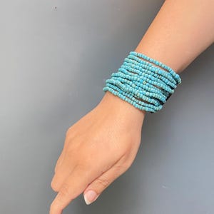 May include: A multi-strand bracelet composed of numerous small, round, turquoise-colored beads. The beads are strung together in multiple rows, creating a wide, layered appearance that wraps around the wrist with a consistent blue hue.
