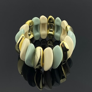 May include: A stretch bracelet composed of alternating curved segments in glossy cream, sage green, and polished gold-tone metal. The individual pieces are arranged in a continuous circular pattern, creating a flexible band with a smooth finish.