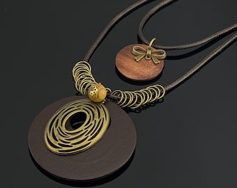 May include: A double-layered necklace featuring a dark brown cord. The lower pendant is a large dark brown wooden circle topped with a brass-toned openwork oval and a small wooden bead. The upper pendant is a smaller wooden circle with a brass bow.