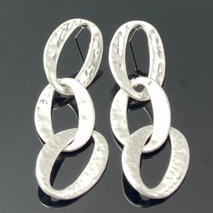 Puede incluir: Pendientes de metal plateado martillado. Cada pendiente presenta tres eslabones ovalados conectados en cadena. Los pendientes tienen una superficie texturizada y un diseño colgante. El fondo es negro.