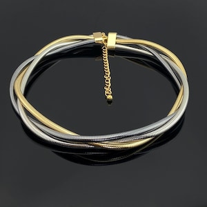 Könnte beinhalten: Eine mehrsträngige Halskette mit ineinander verschlungenen gold- und silberfarbenen Schlangenkettengliedern. Das Design umfasst einen goldfarbenen Metallverschluss und eine verstellbare Verlängerungskette mit einer kleinen Kugel am Ende.