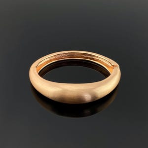 Peut inclure: Un bracelet doré de forme ovale avec une finition en métal brossé. Le bracelet a un design simple et élégant et une fermeture à fermoir. Le bijou est présenté sur une surface noire réfléchissante.