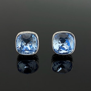 May include: A pair of stud earrings featuring square-cut, light blue faceted crystals set in a polished silver-toned metal frame. The earrings have a rounded square shape with a brilliant, reflective surface and a clear, icy blue colour profile.