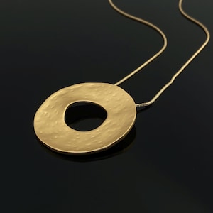 May include: A gold-colored pendant necklace with a large, textured, circular pendant. The pendant has a hole in the center and hangs from a delicate chain. The necklace is displayed against a dark background.