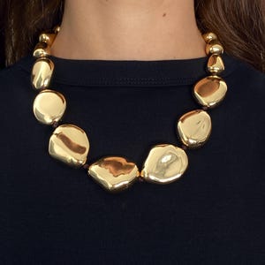 May include: A statement necklace featuring a series of large, irregular, polished gold-tone pebble-shaped beads strung together. The smooth, reflective metallic surfaces of the beads vary in size and shape, creating a continuous, bold circular design.