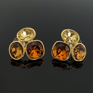 Pode incluir: Um par de brincos de tarracha com três cristais facetados em tons de âmbar e amarelo claro, dispostos em um grupo. Cada brinco é montado em uma estrutura de metal dourado, exibindo uma mistura de pedras de corte redondo e quadrado.