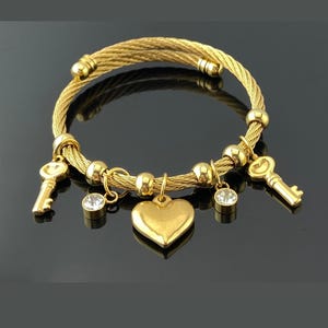 May include: A gold-toned cable wire bracelet featuring a central heart charm, two key charms, and two round clear crystal charms. The bracelet is accented with several smooth gold-toned metal beads spaced along the twisted wire band.