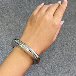 May include: A thick, polished silver-tone metal bangle bracelet with a smooth, rounded, and reflective surface. The bracelet features a substantial, chunky design that encircles the wrist, showcasing a high-shine finish that captures light.