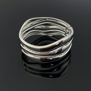 Pode incluir: Uma pulseira de punho articulada em tom prateado com um design de várias camadas, apresentando três bandas distintas, lisas e curvas que se sobrepõem para criar uma estrutura aberta e fluida. O metal tem um acabamento polido e reflexivo.