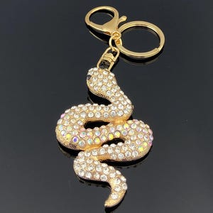 Pode incluir: Um porta-chaves em metal dourado com um pendente em forma de serpente enrolada. A serpente está coberta de strass transparentes e iridescentes que captam a luz. O porta-chaves inclui um anel circular e um fecho lagosta para fixação segura.