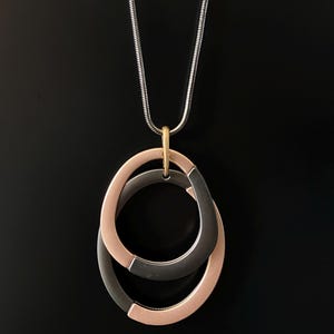 Könnte beinhalten: Eine Halskette mit einer silberfarbenen Schlangenkette und einem Anhänger aus zwei ineinandergreifenden, asymmetrischen Ringen. Ein Ring ist matt roségoldfarben, der andere dunkel anthrazitgrau. Eine kleine goldfarbene Öse verbindet die Ringe.