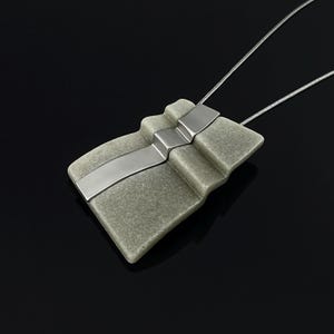 May include: A pendant necklace featuring a rectangular, textured sage green stone element with a stepped, layered surface. A smooth, polished silver-toned metal strip runs diagonally across the centre of the stone, attached to a thin silver-toned chain.