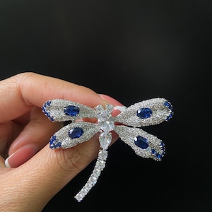 May include: A dragonfly-shaped brooch featuring a silver-toned metal frame encrusted with numerous small clear crystals. The wings are accented with four oval-shaped deep blue stones, while the body and head are set with clear pear-shaped crystals.