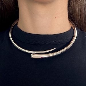 Könnte beinhalten: Eine polierte silberfarbene Metall-Halskette im offenen Design. Die Kette besteht aus einem starren, geschwungenen Band, das sich vorne zu abgerundeten Spitzen verjüngt und so einen überlappenden Effekt um den Hals erzeugt.