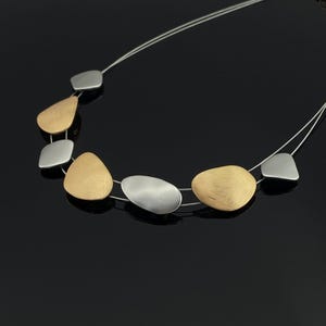 May include: A silver necklace featuring a series of irregularly shaped pendants in gold and silver tones. The pendants are strung on a thin, delicate silver wire, creating a modern and minimalist design. The necklace is displayed against a black background.