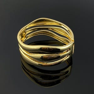 Könnte beinhalten: Ein goldfarbener Klapparmreif mit mehrsträngigem Design, bestehend aus drei glatten, geschwungenen und polierten Metallbändern, die an einem sicheren Verschlussmechanismus zusammenlaufen und eine fließende, offene Kreisform bilden.