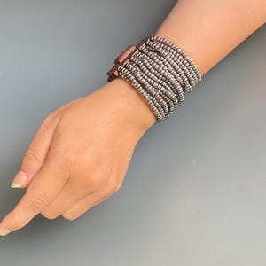 May include: A multi-strand bracelet with small, silver-coloured beads. The bracelet is fastened with a rectangular, dark brown clasp. The jewellery is worn on a wrist against a grey background.