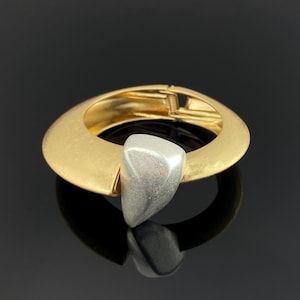 Puede incluir: Una pulsera rígida articulada con una banda ancha de tono dorado mate y silueta curva y lisa. Un prominente acento triangular de tono plateado texturizado está fijado en la parte frontal de la banda, creando un contraste de color y acabado.