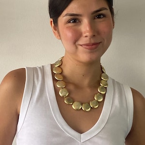 May include: A statement necklace featuring a series of round, metallic gold-toned discs linked together in a single strand. The discs have a textured, slightly irregular surface finish and are worn around the neck over a plain white sleeveless top.