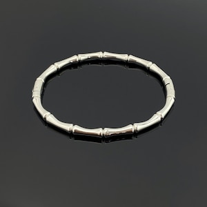 Puede incluir: Una pulsera de tono plateado pulido con un diseño segmentado que imita la apariencia de tallos de bambú. El brazalete circular está construido a partir de múltiples eslabones metálicos conectados con un acabado liso y reflectante.