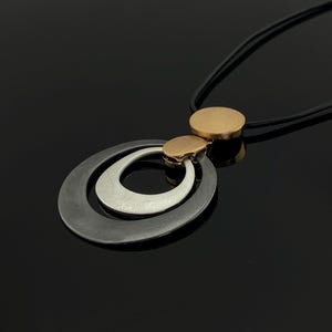 Könnte beinhalten: Eine moderne Halskette mit einem schwarzen ovalen Anhänger, einem silbernen Innenoval und einem goldfarbenen Akzent. Die Halskette hat eine schwarze Kordel und einen goldfarbenen kreisförmigen Anhänger oben. Der Schmuck ist vor schwarzem Hintergrund platziert.