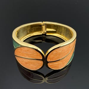 Op de afbeelding: Een goudkleurige scharnierende armband met een bladvormig ontwerp, voorzien van gladde, gemarmerde perzikkleurige emaille inlays en groene emaille accenten aan de zijkanten. De binnenzijde van de metalen band heeft een textuur met arceringen.