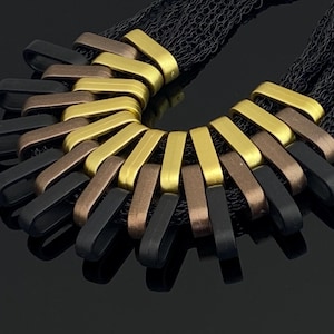 Könnte beinhalten: Eine auffällige Halskette mit einer schwarzen Mesh-Kette und einer Reihe von gebogenen, rechteckigen Gliedern in Gold, Bronze und Schwarz. Die Glieder sind in einem halbkreisförmigen Muster angeordnet und schaffen ein kühnes und modernes Design. Die Halskette wird vor einem dunklen Hintergrund präsentiert.