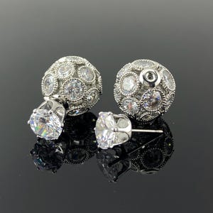 Op de afbeelding: Twee zilverkleurige, dubbelzijdige oorstekers. Elke oorbel heeft een grote, heldere, ronde edelsteen aan de voorkant en een kleinere, getextureerde, ronde achterkant bezet met kleinere heldere edelstenen. Op een reflecterend zwart oppervlak.