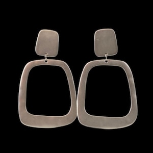 Puede incluir: Un par de pendientes plateados con un diseño geométrico. Cada pendiente presenta una pieza superior rectangular conectada a una forma cuadrada abierta más grande. Los pendientes cuelgan sobre un fondo negro.