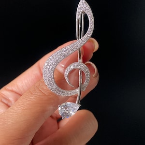 Peut inclure: Une broche en forme de clé de sol argentée, incrustée de nombreuses petites pierres claires et scintillantes. Le motif musical présente une pierre unique plus grande en forme de poire à la base, créant un accessoire métallique brillant.