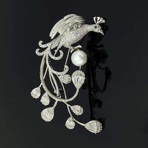 May include: A silver-toned brooch in the shape of a peacock, adorned with sparkling clear crystals. The bird has a pearl and a small green gem. The brooch has a floral design with crystal accents, creating an elegant and detailed accessory.