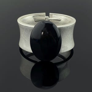 Könnte beinhalten: Ein breiter Armreif mit einem gebürsteten, silberfarbenen Metallband in matter Optik und einem großen, glatten, ovalen schwarzen Stein in der Mitte. Das Armband verfügt über einen Scharnierverschluss, der an der Oberseite des Bandes sichtbar ist.