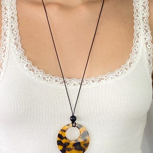 May include: A long necklace featuring a circular pendant with a brown and black tortoiseshell pattern. The pendant hangs from a thin black cord, which is accented by a single small black bead positioned directly above the circular charm.