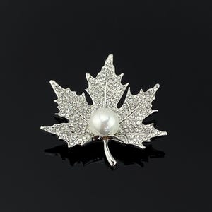 May include: A silver-toned maple leaf brooch featuring a polished white faux pearl at the centre. The entire surface of the leaf is encrusted with numerous small, clear, sparkling rhinestones, creating a textured and reflective appearance.