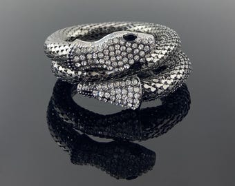 May include: A coiled snake bracelet in a dark metallic finish featuring a textured scale pattern. The head and tail are encrusted with clear, sparkling rhinestones, and the eyes are accented with small, dark, oval-shaped stones.