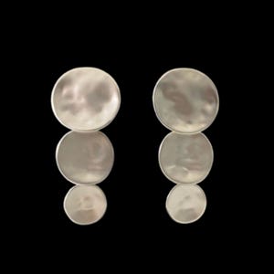 May include: A pair of silver-toned, layered disc earrings. Each earring features three graduated, hammered-texture circles suspended vertically. The earrings are set against a black background, highlighting their metallic sheen and design.