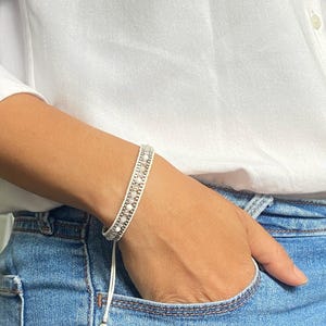 Puede incluir: Una pulsera envolvente blanca con pequeñas cuentas de color claro y detalles plateados. La pulsera se lleva en la muñeca, con un cierre de cordón blanco. La persona lleva una camisa blanca y vaqueros azules.