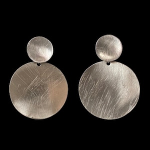 Op de afbeelding: Een paar zilverkleurige oorbellen met een ontwerp van twee schijven. Elke oorbel bestaat uit een kleinere ronde knop verbonden met een grotere ronde hanger. Beide schijven hebben een geborstelde, getextureerde afwerking met fijne lijnen.