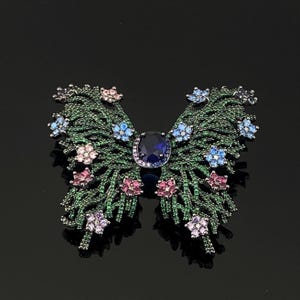 May include: A butterfly-shaped brooch featuring a dark metal frame encrusted with small green stones. The wings are decorated with clusters of light blue, pink, and purple floral-shaped stones, and a large oval dark blue stone sits at the centre.